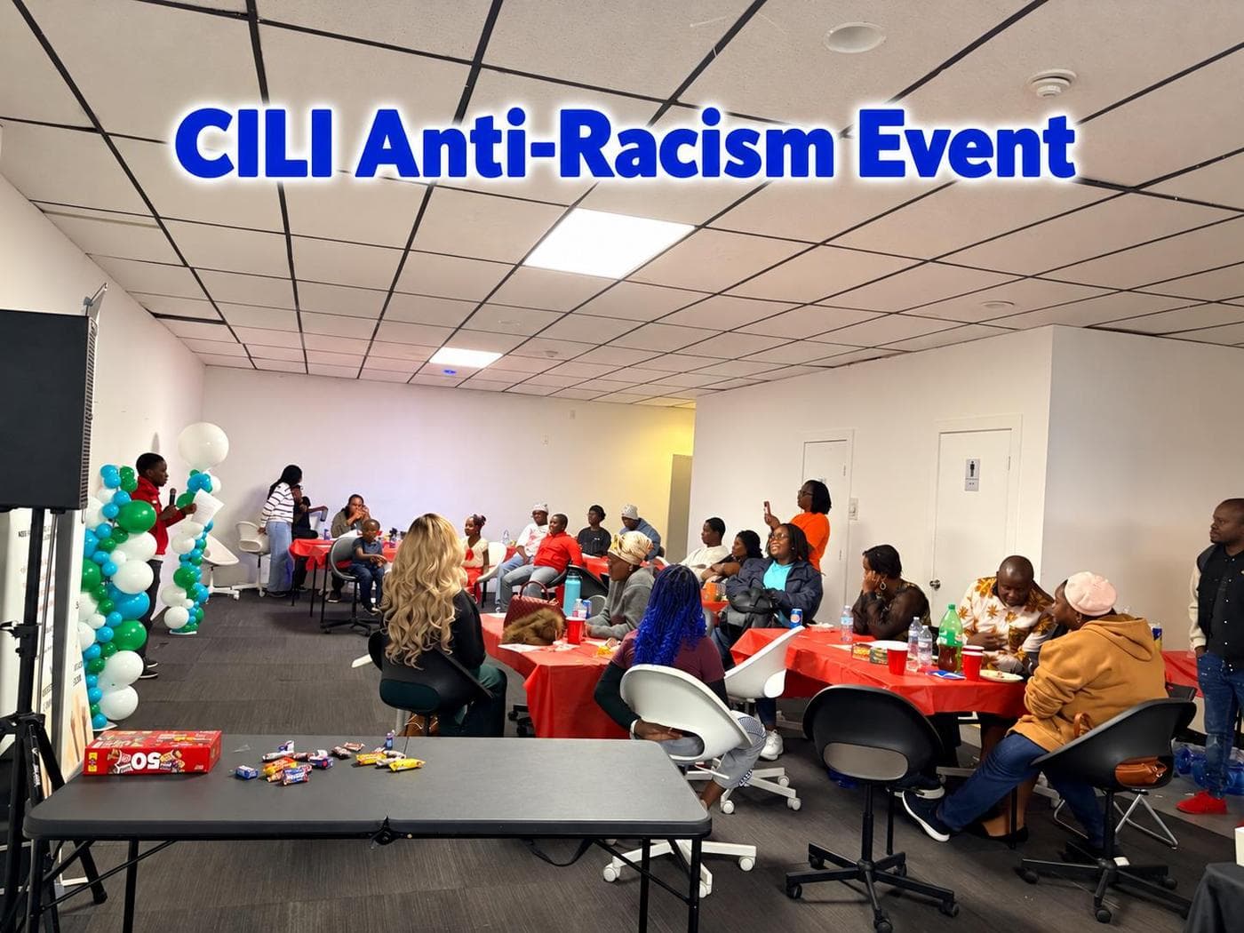 Community members seated at tables during the CILI Anti-Racism Event, with teal and white balloons and a CILI Anti-Racism Event banner on the wall.